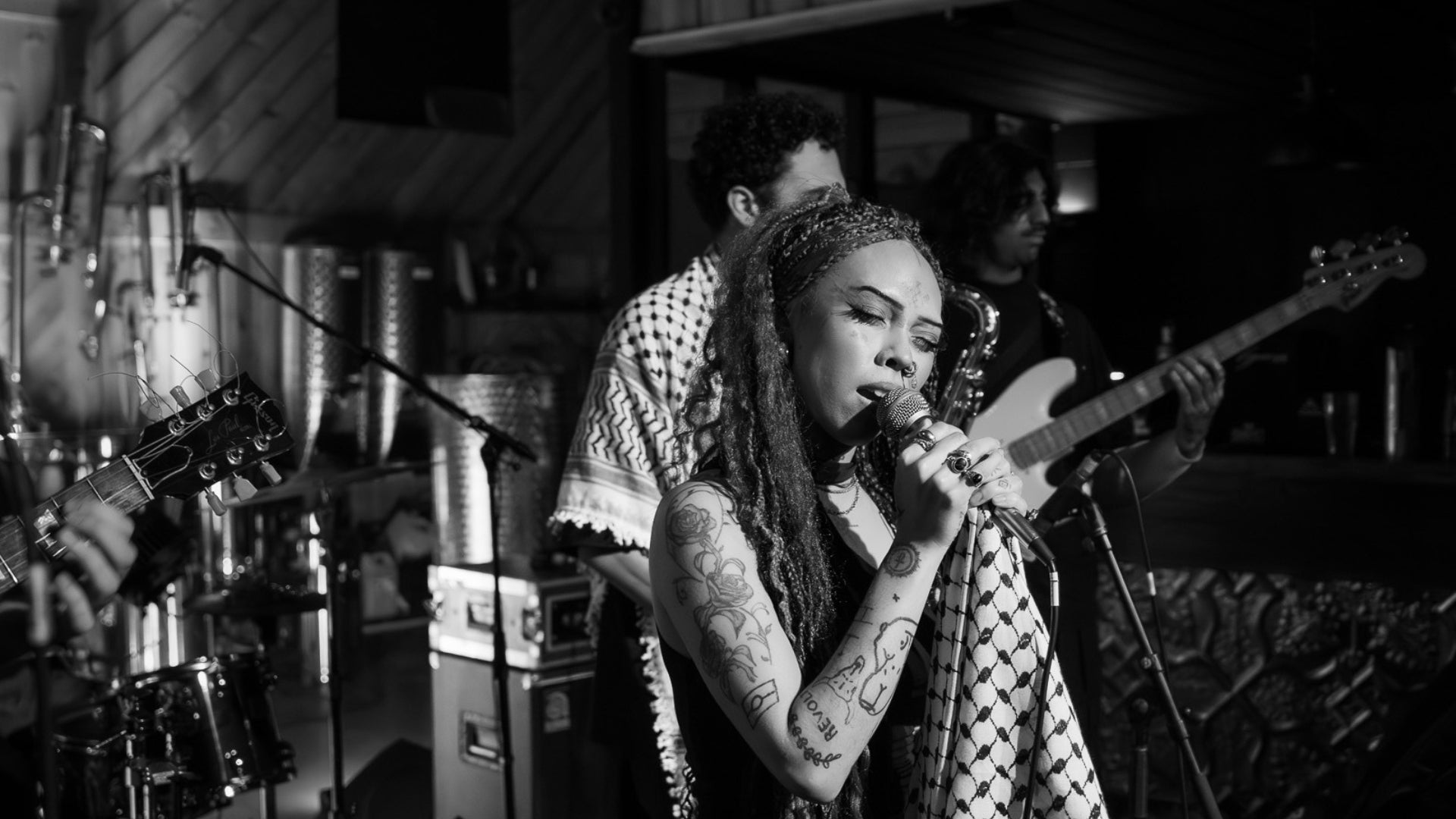 Black and white photo of person singing into microphone. The person has tattoos on their arm and are holding a towel. A guitarist and other band members can be seen in the background. 