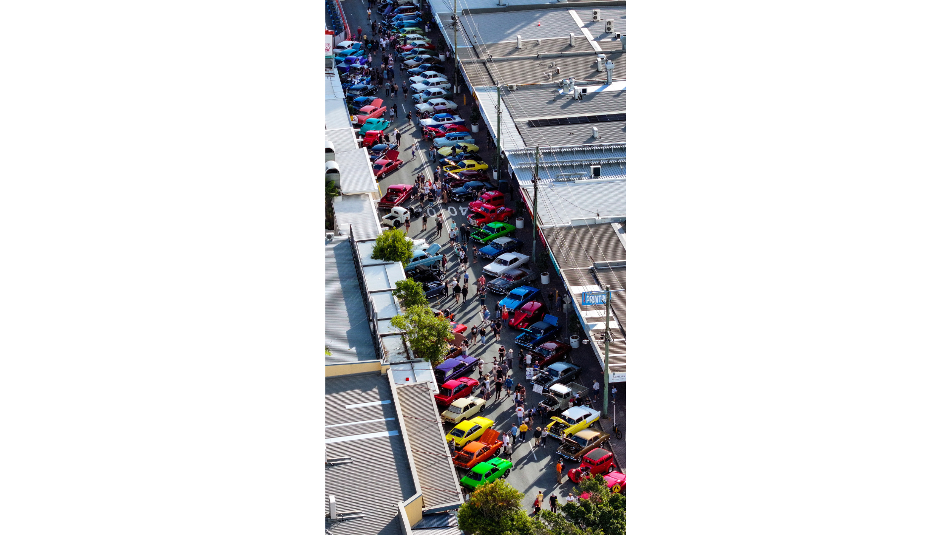 An aerial view of lots of colourful classic and custom cars parked on both sides of the street - with buildings either side.