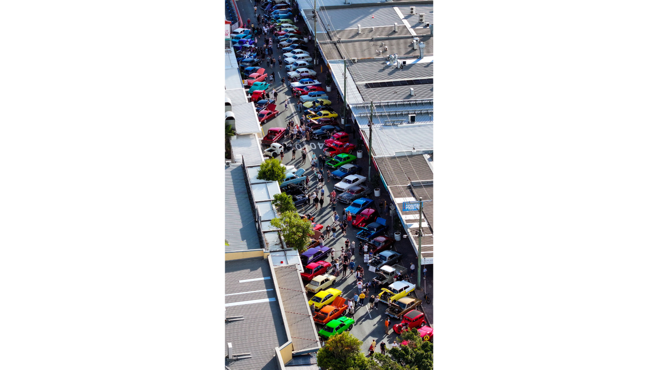An aerial view of lots of colourful classic and custom cars parked on both sides of the street - with buildings either side.