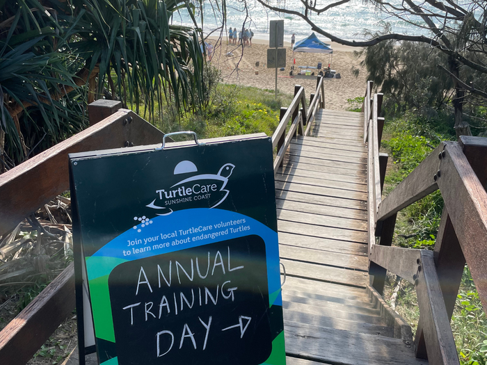 A sign on a timber staircase leading down to a timber walkway. The sign features the TurtleCare logo and the words "annual training day." In the background, a beach scene with a blue marquee and people standing underneath it. Beyond that, the blue ocean sparkles under the sun's reflection.