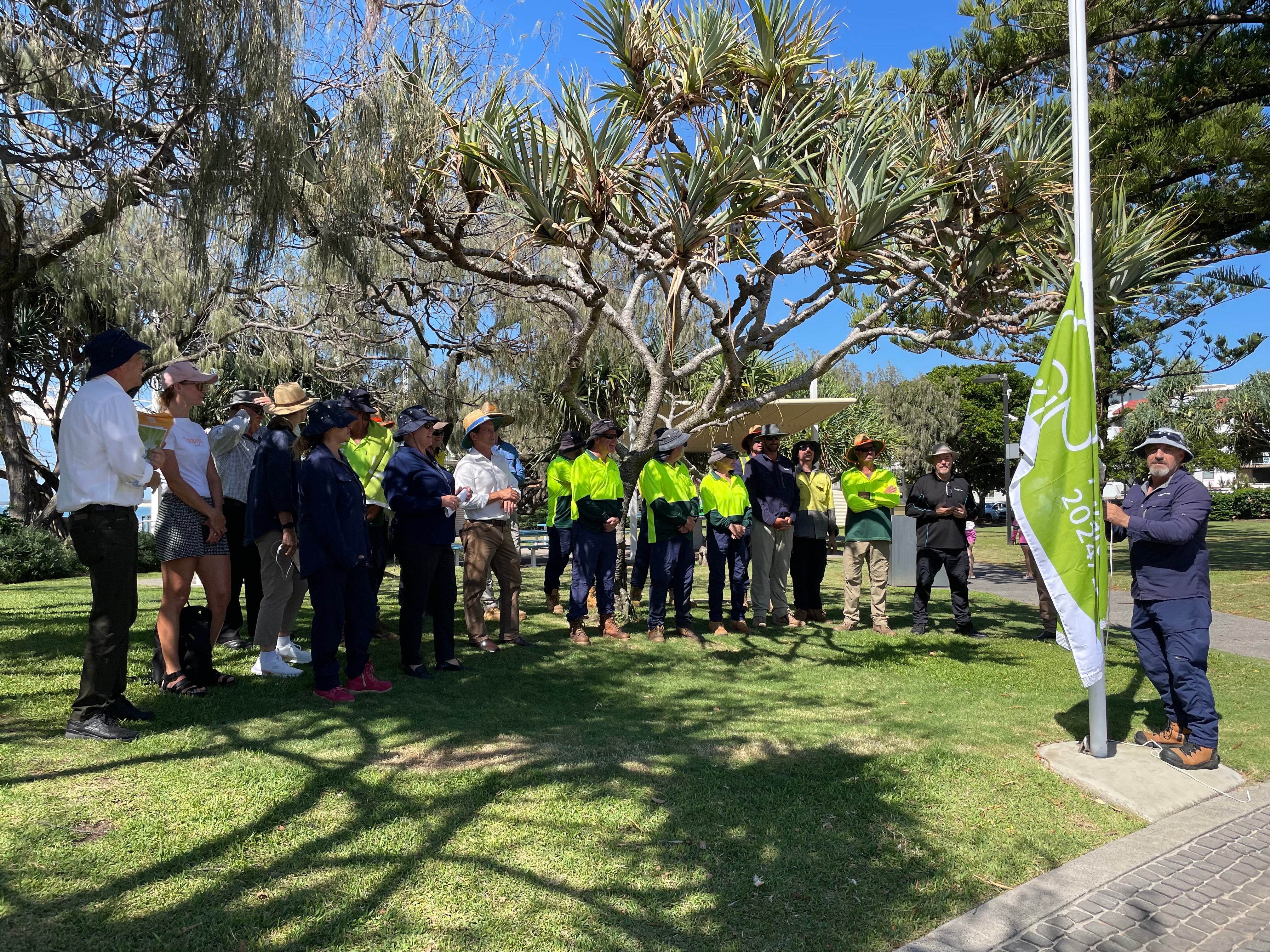 Group of people standing under green pandana trees on green grassland. A person adding to the flag pole the Green Flag Award 2024 to 2025. 