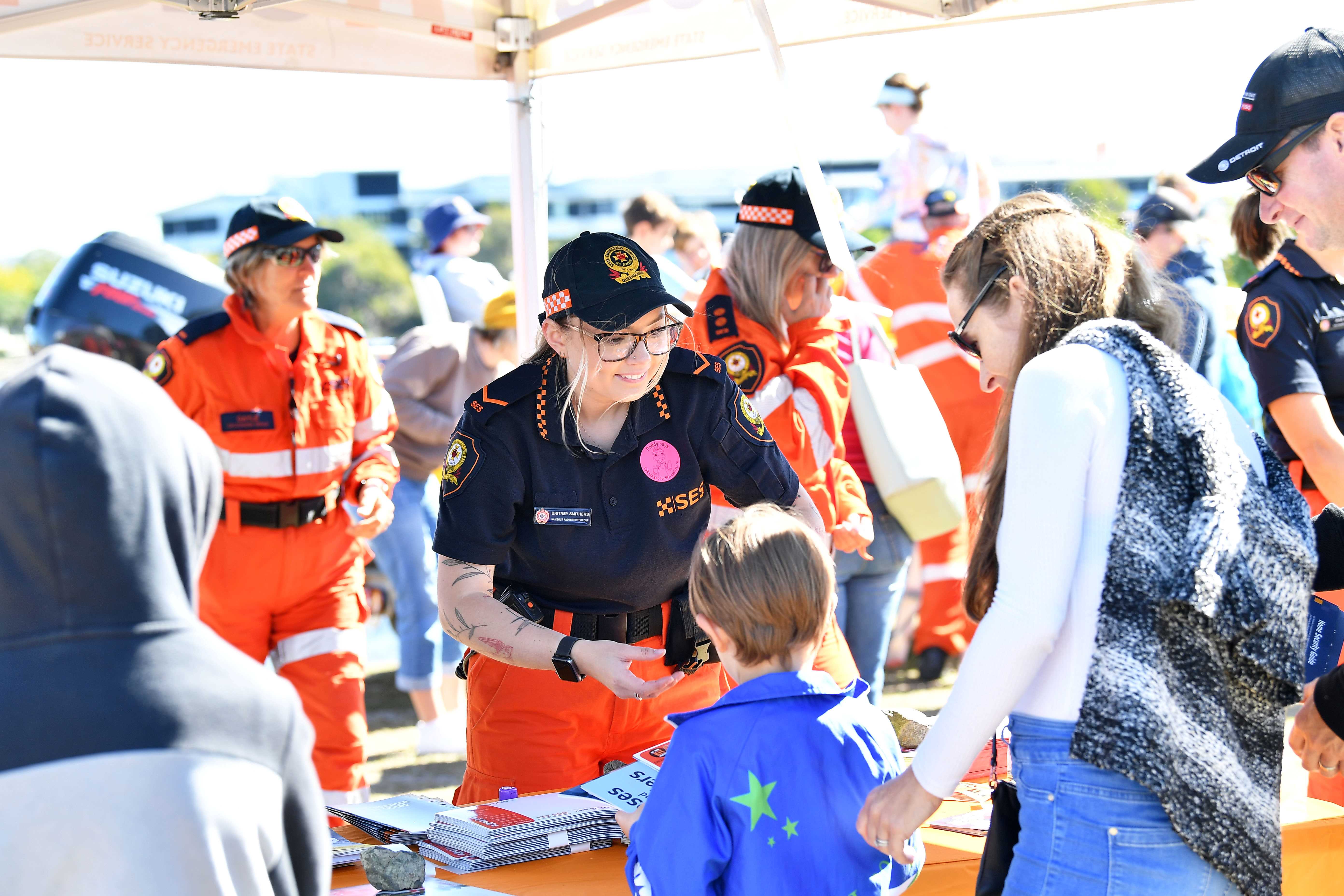 A smiling SES volunteer stands behind a stall surrounded by a crowd. She is handing something to a small boy with his mother.