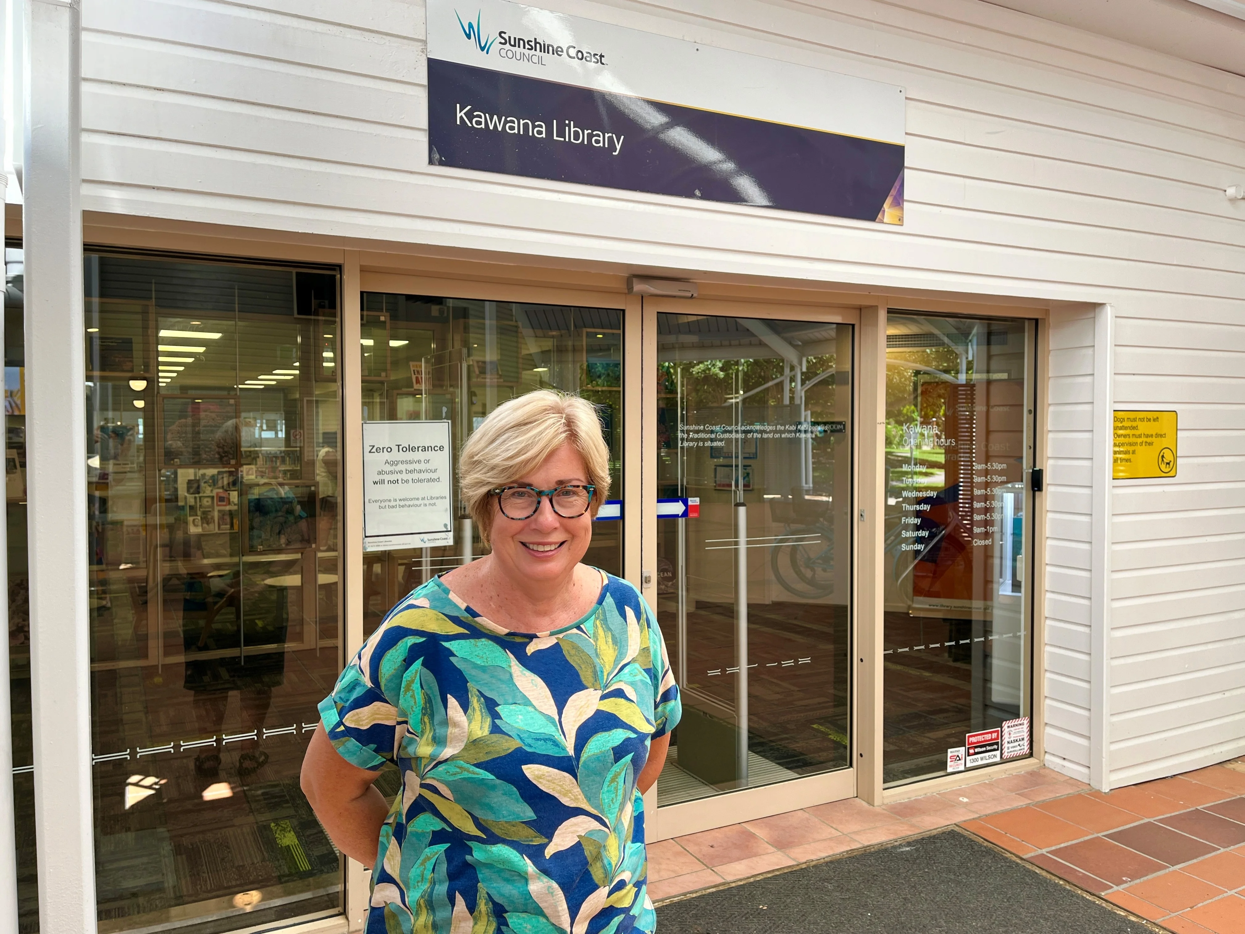 Volunteer Lyn Gavin standing outside the Kawana Library, hands folded behind her back, looking at the camera smiling. 