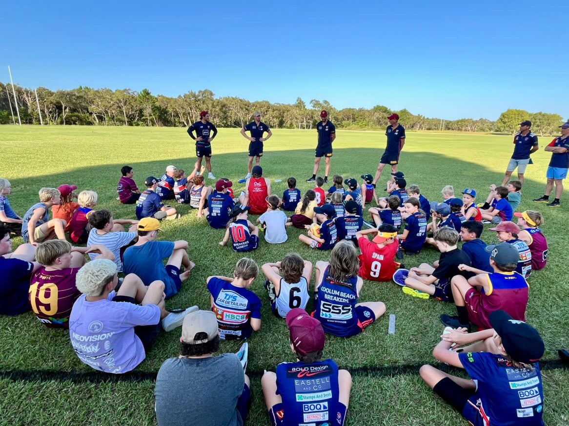 AFL players standing in front of a group of budding young players sitting on the ground