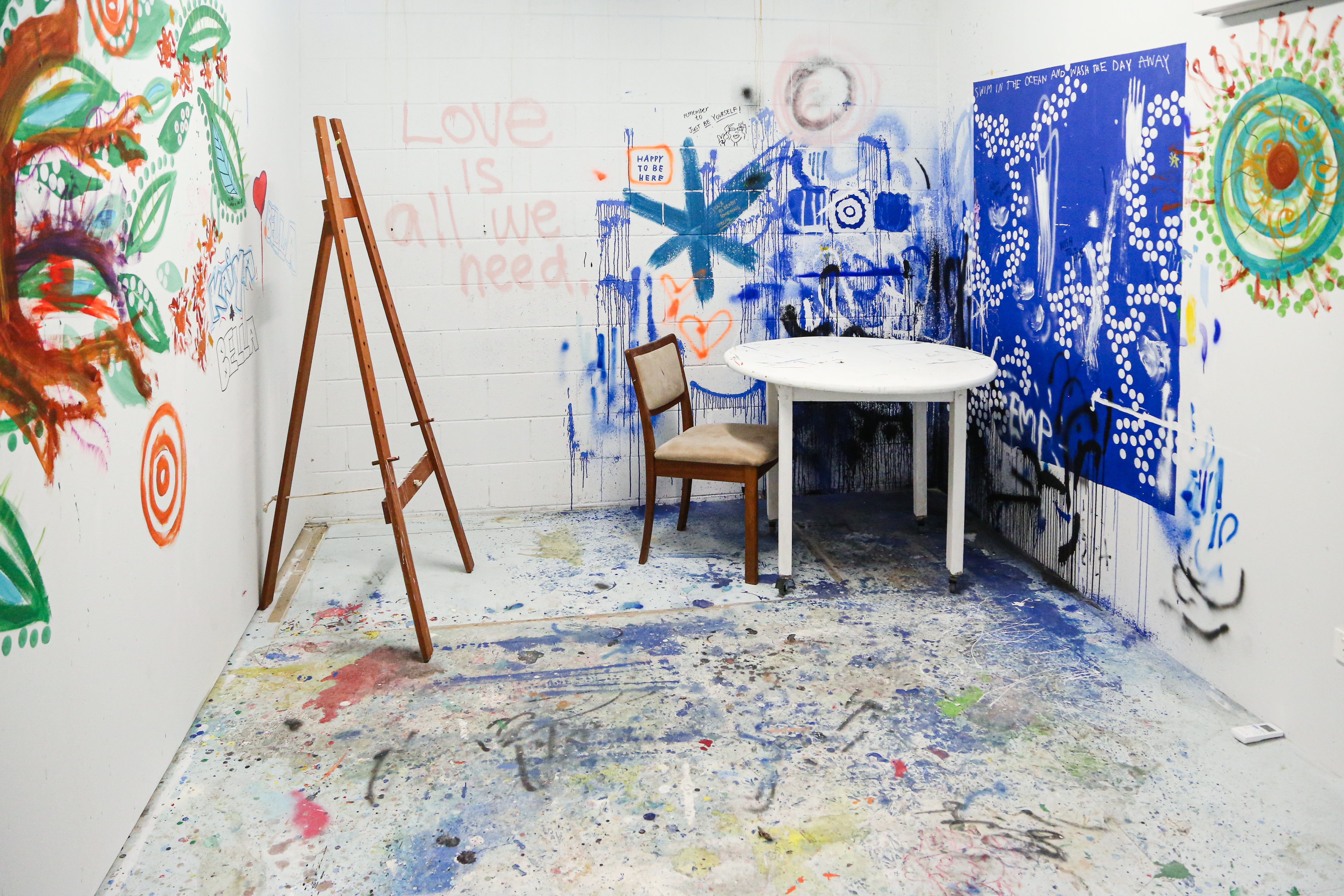 Artist space with paint on the floor, an easel, table and chair, and artworks painted directly onto the white walls