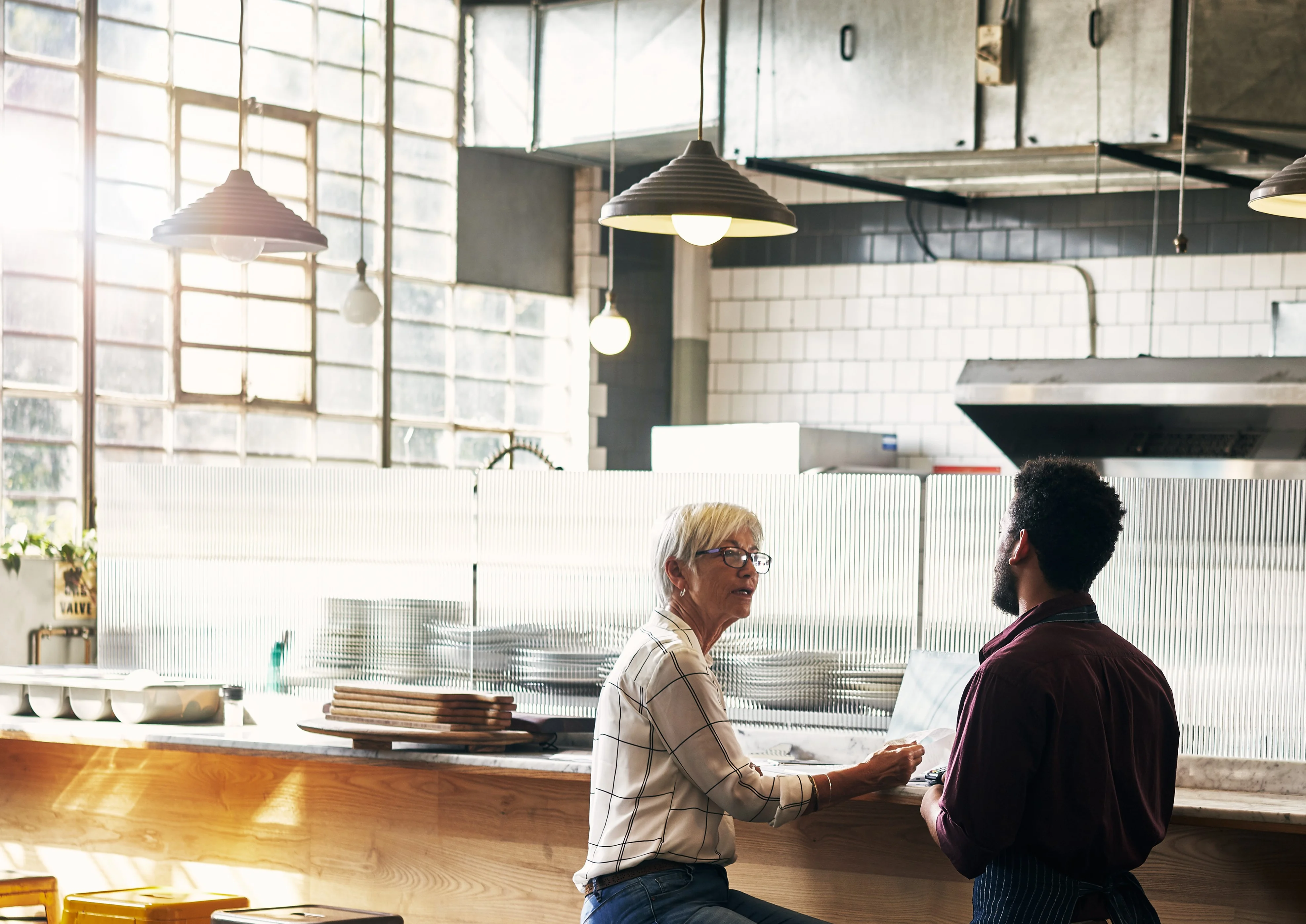 A mature woman sits in a closed restaurant holding paperwork while speaking to a with a young waiter.