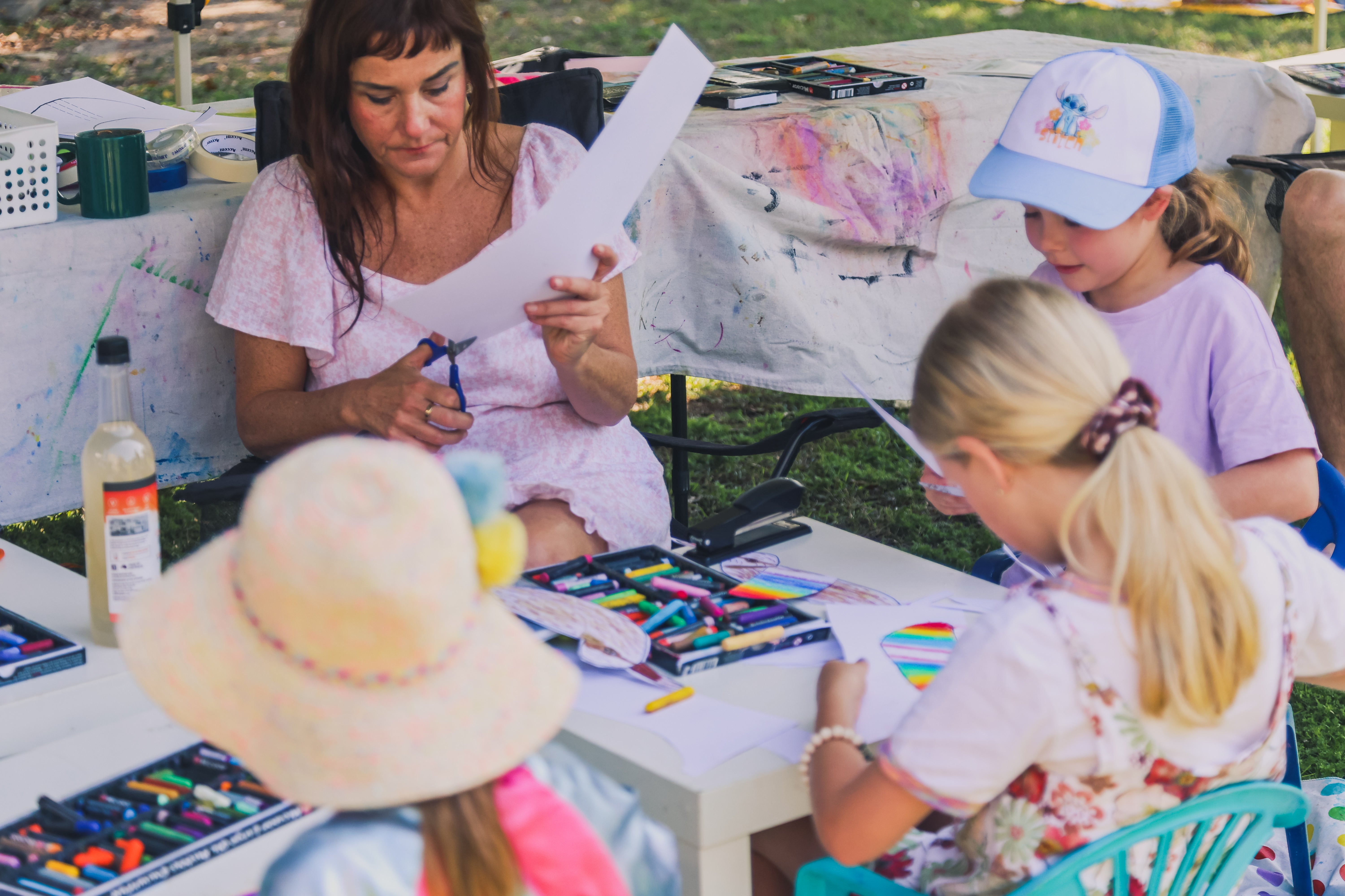 "People enjoying a shorebird festival on a sunny day in a park, with children painting and getting face paint, adults chatting and learning about migratory birds, and various activity stalls visible in the background."