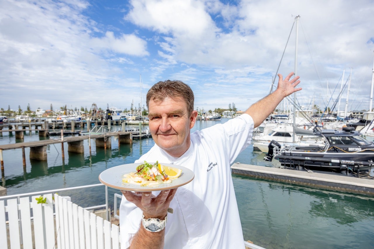 Murray Paterson Executive Chef at Pier 33 is holding a plate of food in front with his other arm up in the air as if to showcase the marina and boats behind their venue.