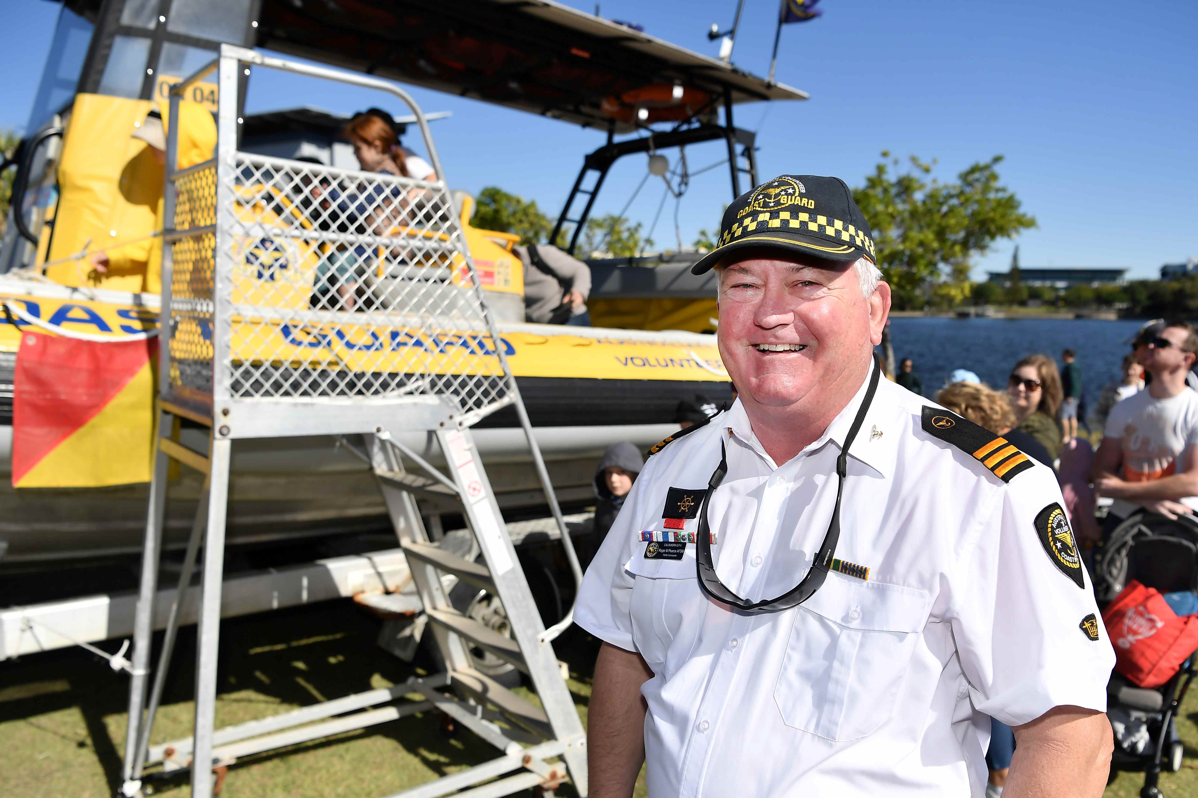 An older man in uniform stands in front of a Coast Guard boat with children and their parents in the background.