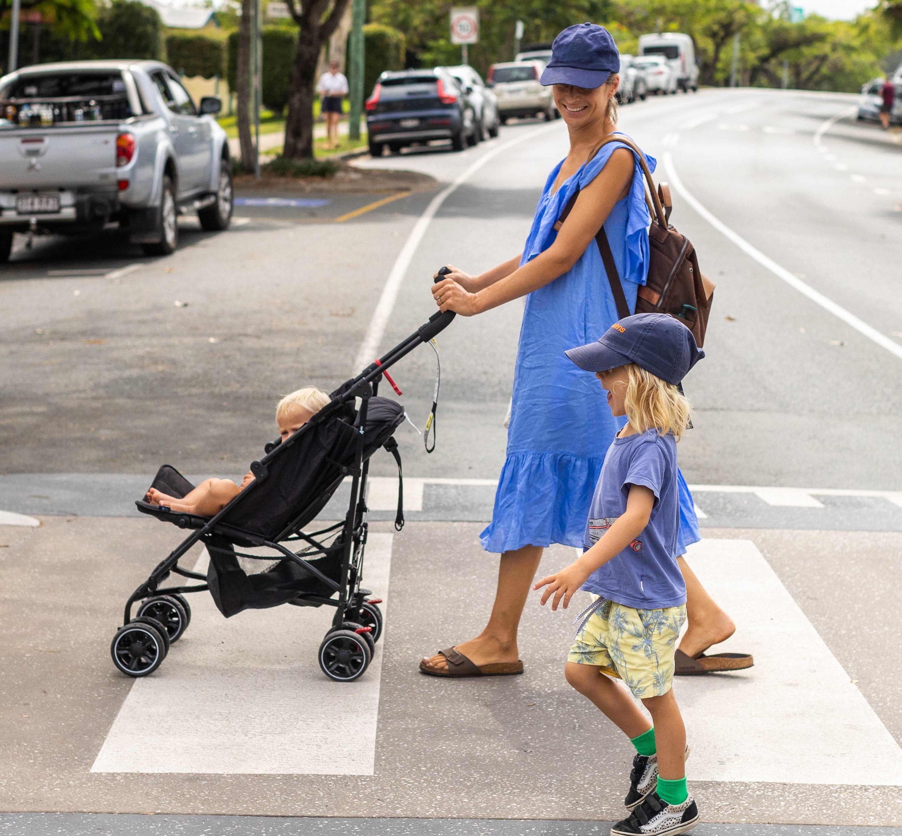 A little boy walks across a zebra crossing with his Mum who is pushing a troller with a baby.