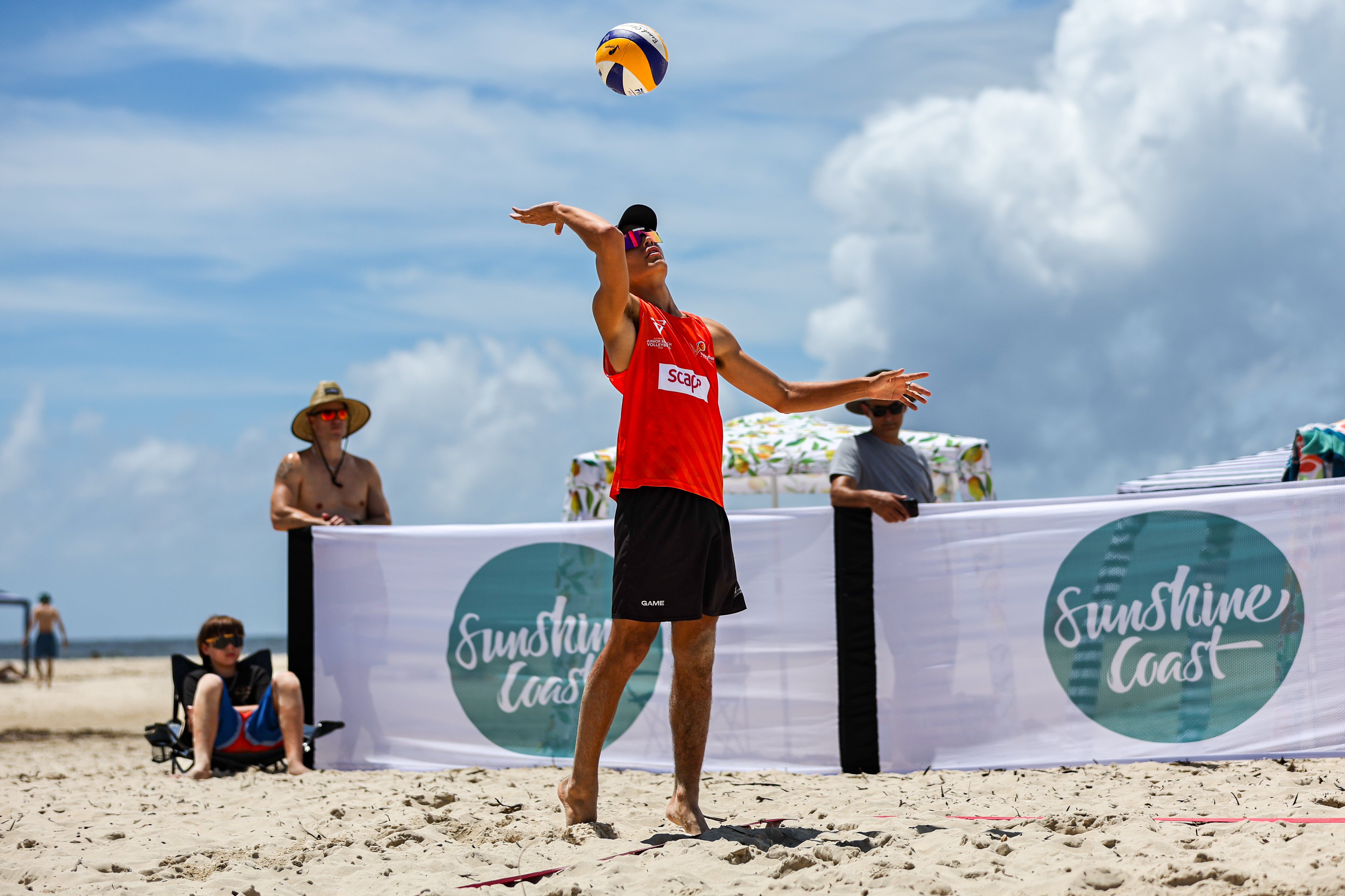 A beach volleyball player on the beach with the ball in the air with arms ready to spike or serve. Background is sky and clouds, and destination signage saying Sunshine Coast.