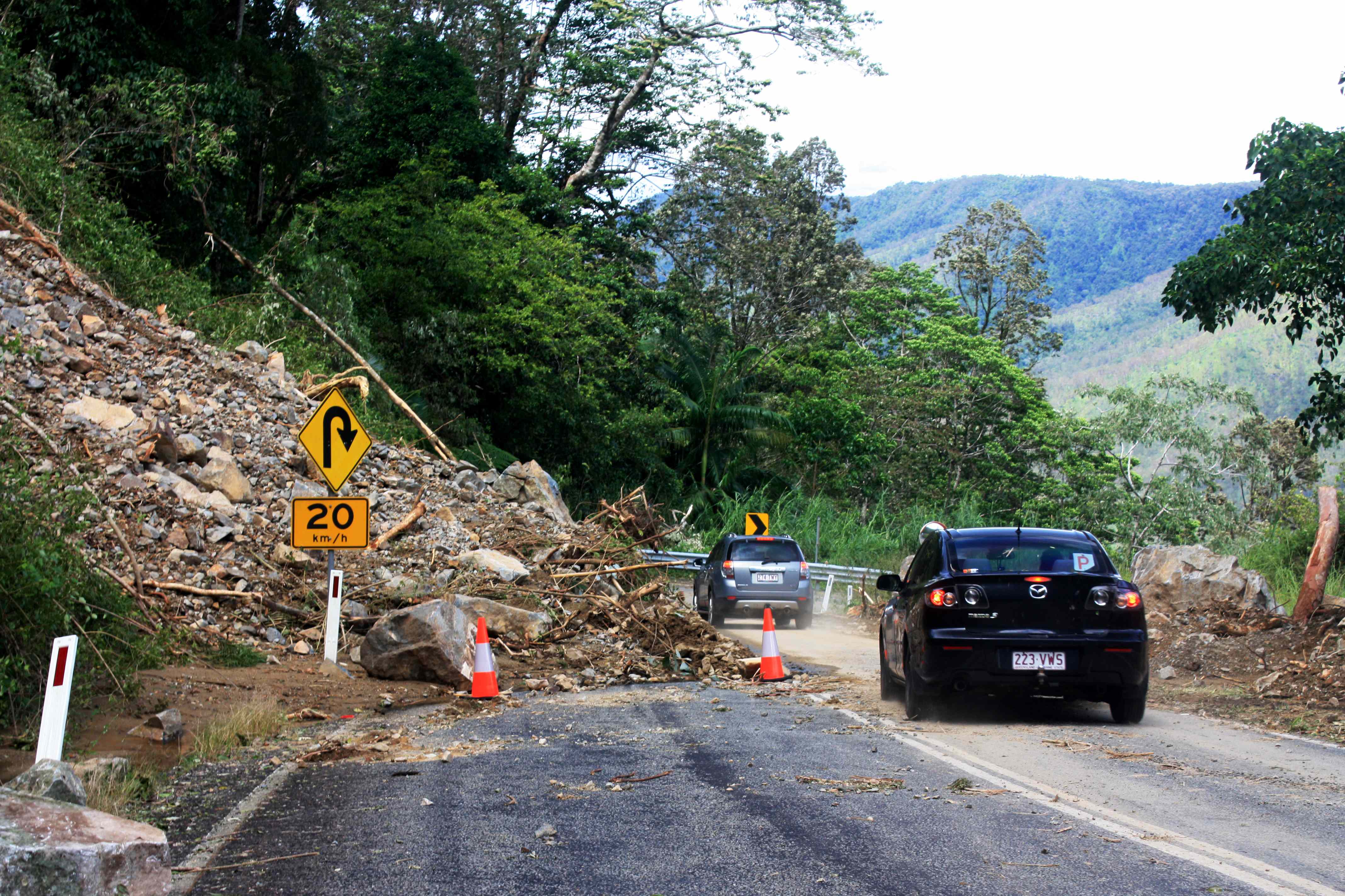 A line of cars cautiously drives past fallen debris from a cliff on a hinterland road.