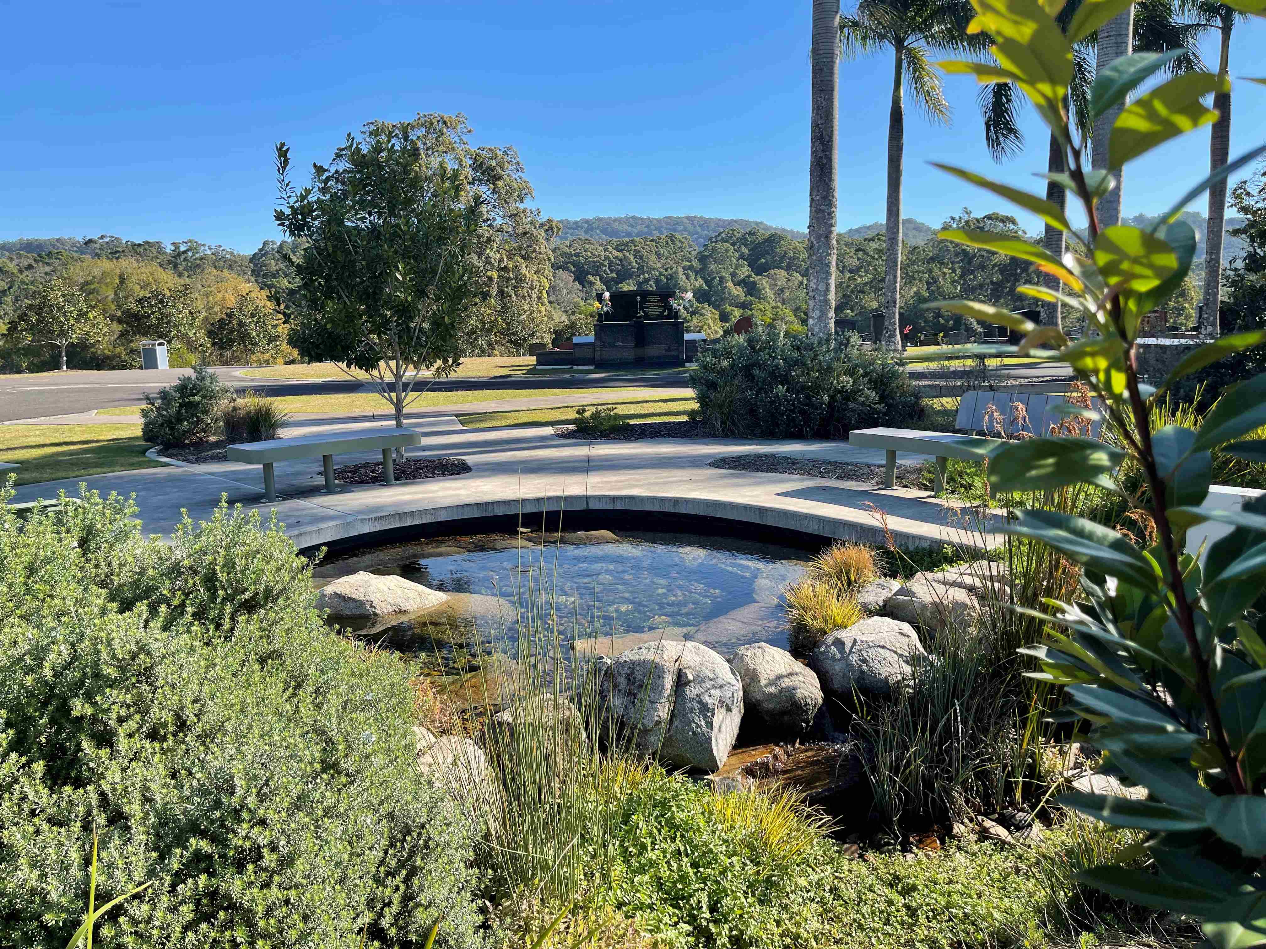 A view of the reflection pond with a seat, surrounded by trees at Kulagoor Cemetery.