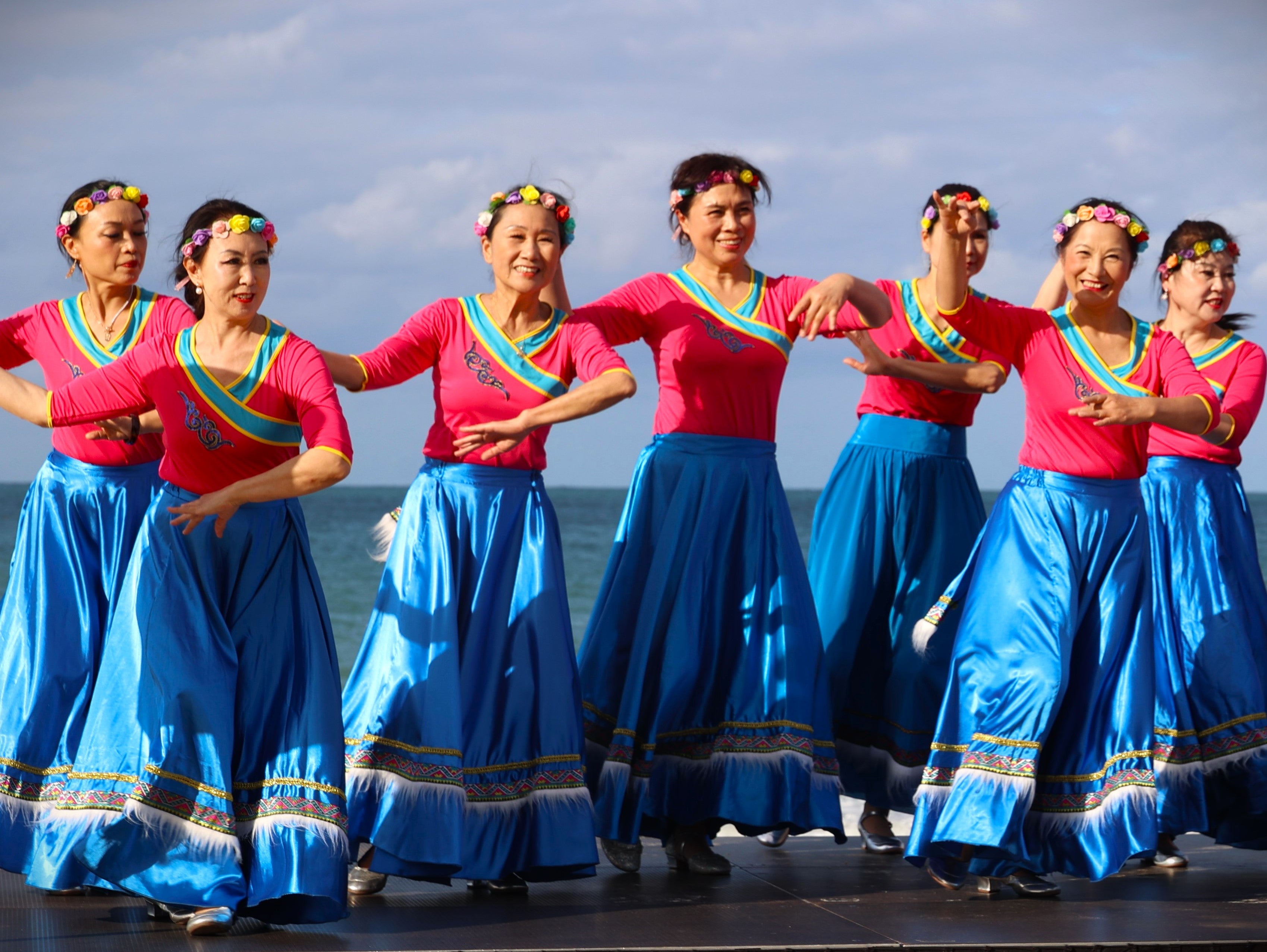 Seven women wearing colourful, traditional Chinese costumes dance on an outdoor stage together, in front of the ocean.