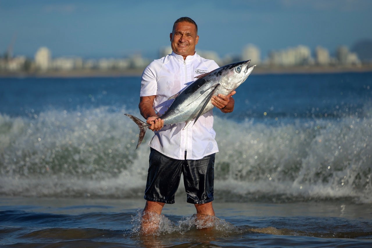 Peter Kuruvita is holding a huge fish while standing in shallow waters and a wave crashing behind him.