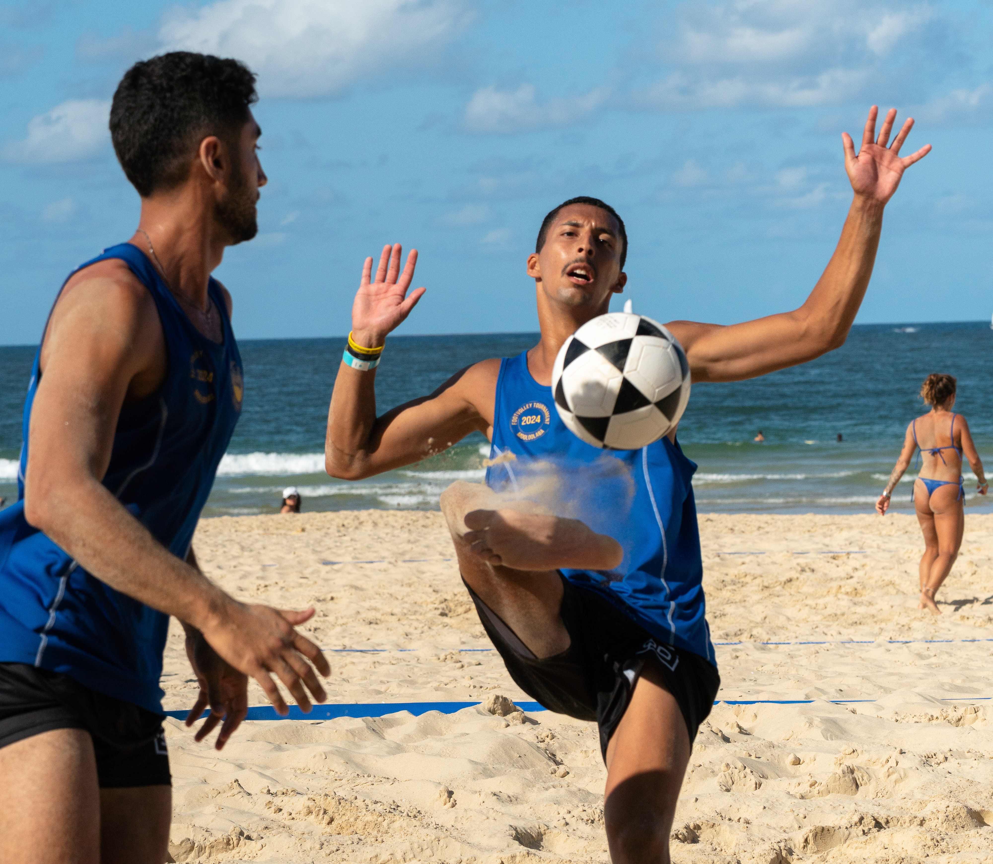 Two men wearing blue singlets are on the beach beside a volleyball net. The man on the right holds his hands up and kicks the ball with his foot.