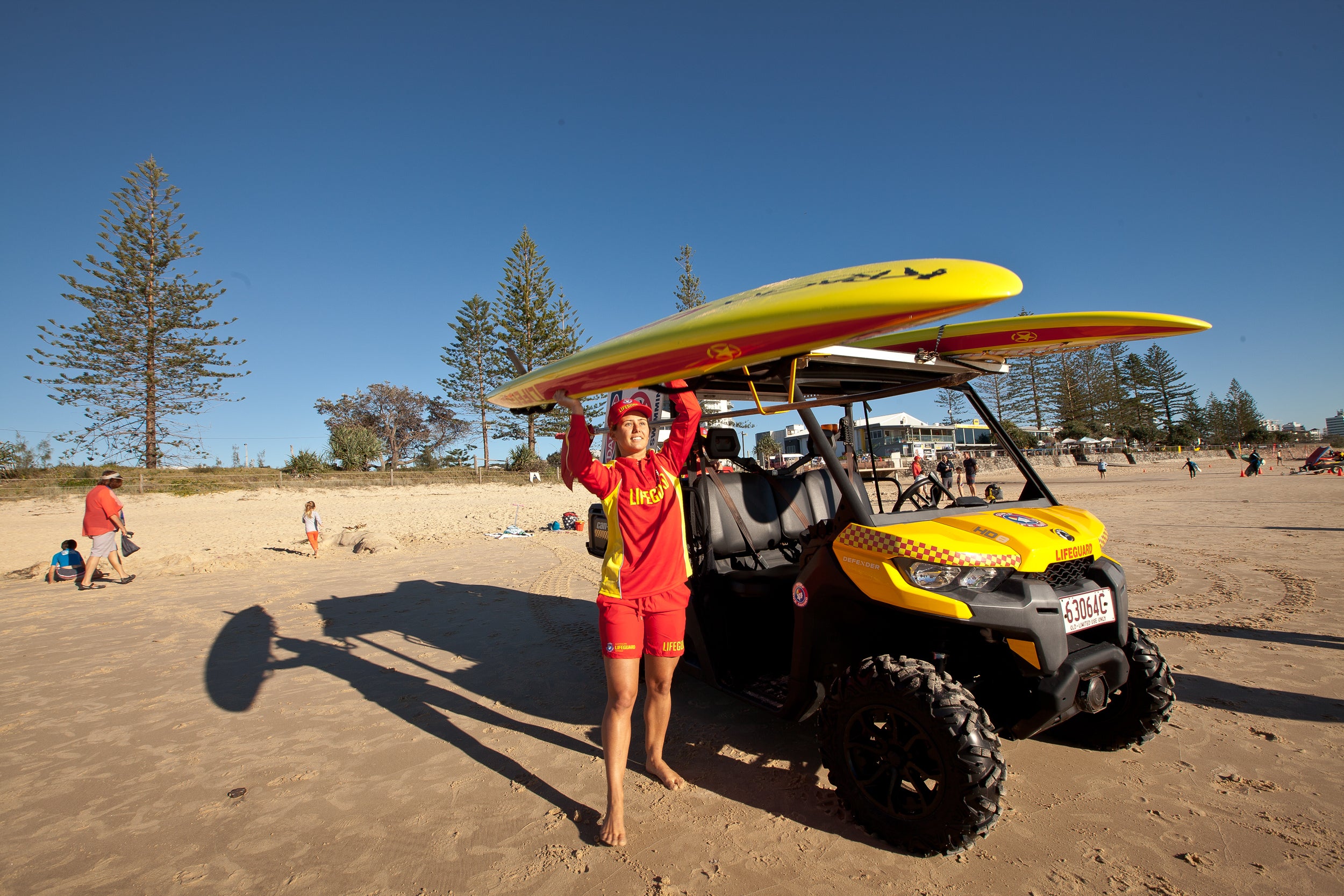 On a beach landscape with blue skies and trees on the horizon, a lifeguard dressed in a red uniform is holding up a yellow surf ski (rescue board) removing it from the roof of their beach patrol vehicle - an ATV (all terrain vehicle).