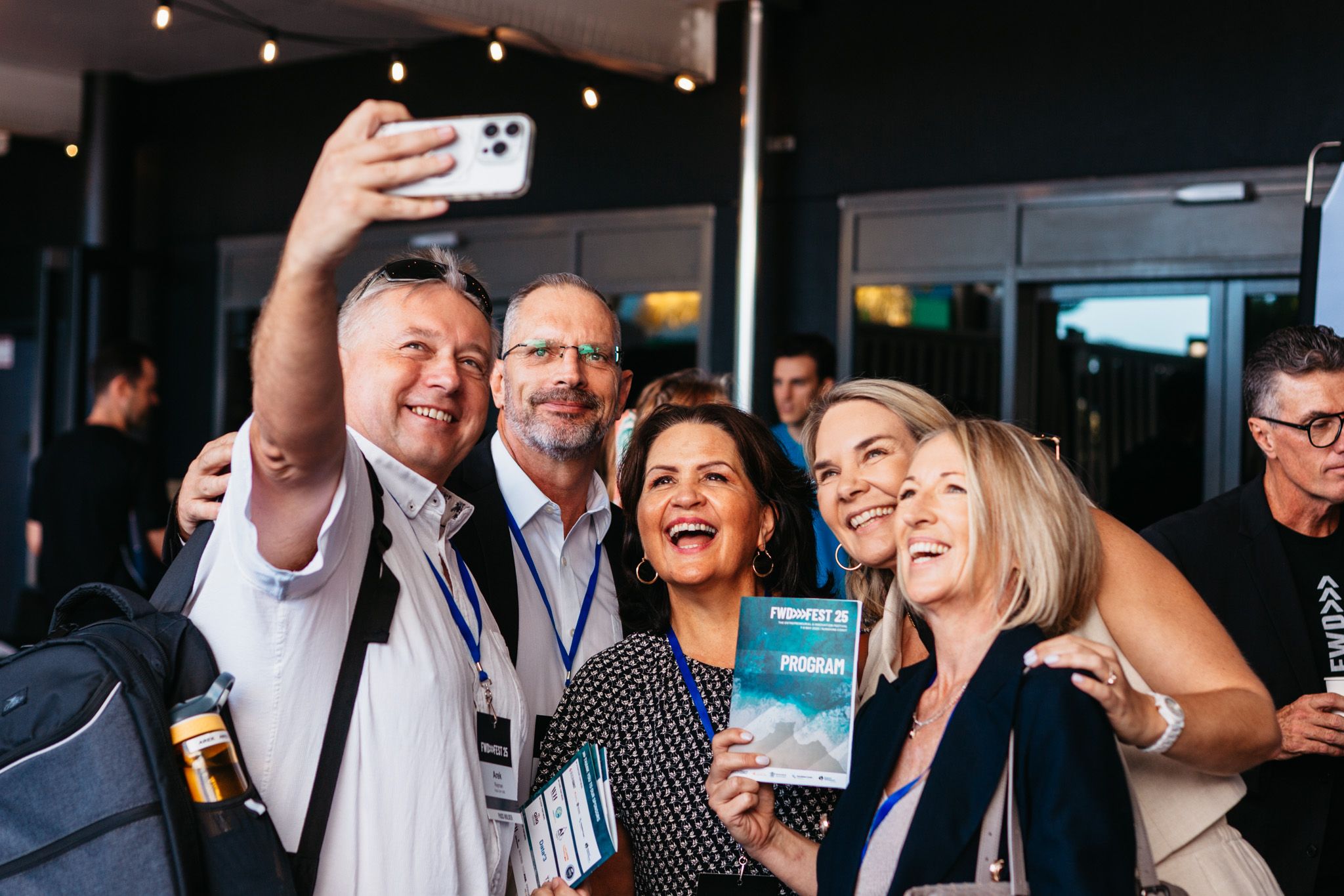 A group of people smile at a phone camera while holding the event catalogue.