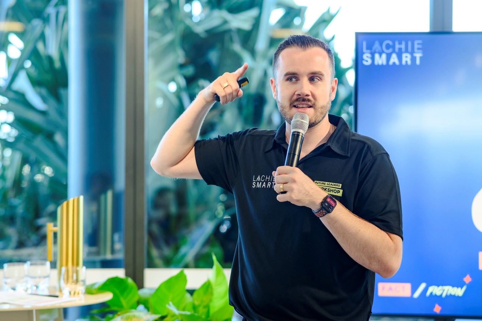 Mr Lachie Smart speaking at a workshop; holding a microphone in one hand and his other hand up in the air - almost pointing to his head - like an idea expression.