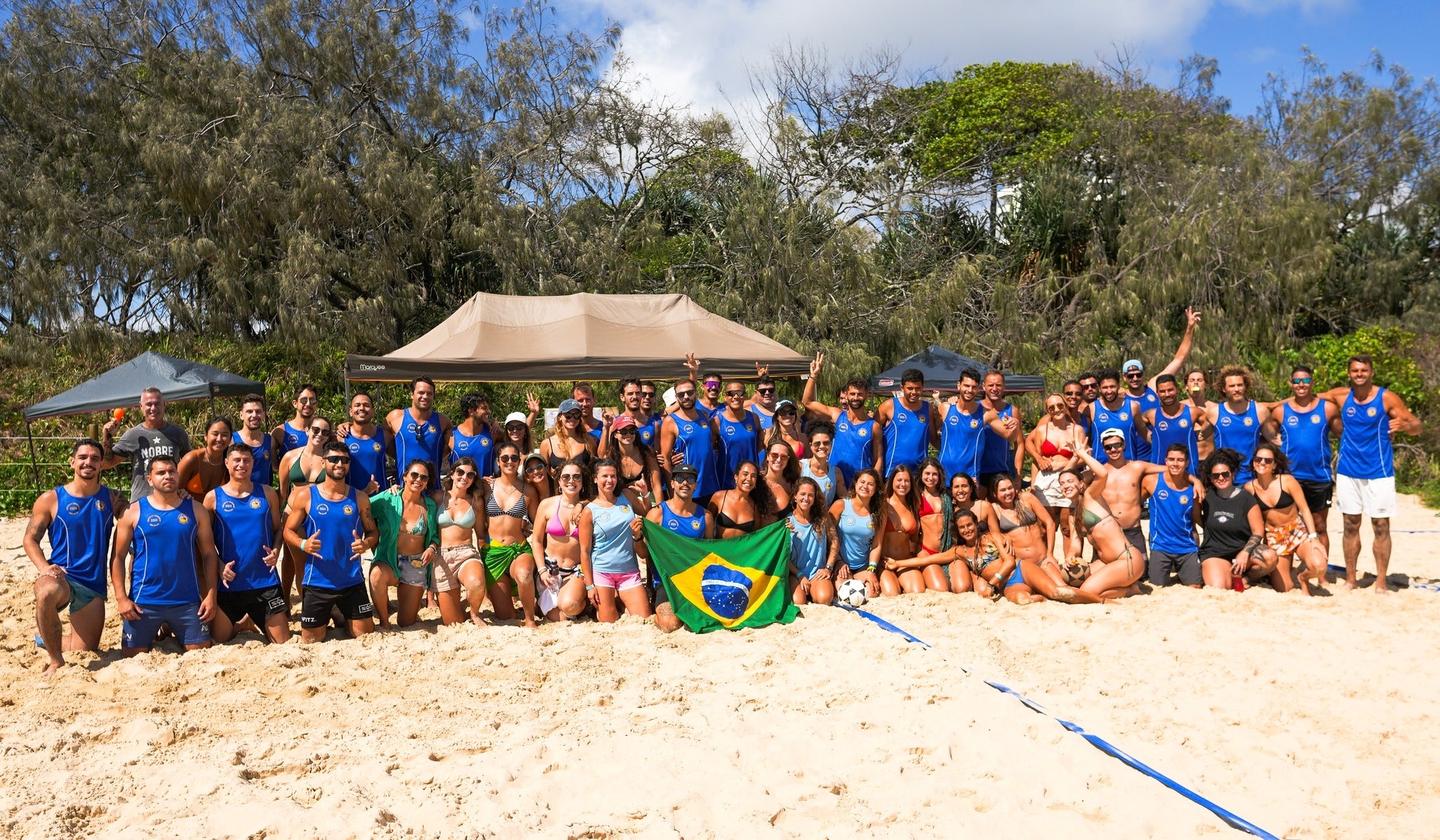 A large group of happy people are gathered together in front of a marquee on a beach, with a Brazilian flag held up in the front. Many wear blue sport singlets and swimsuits. 