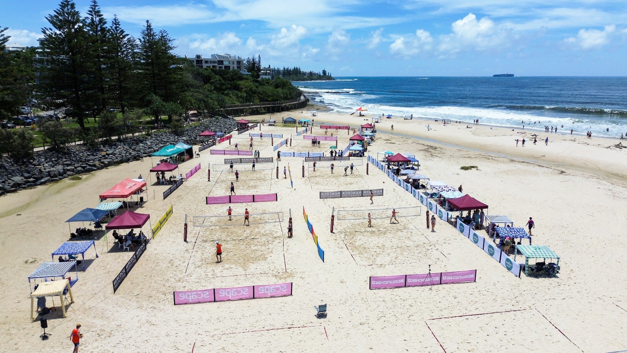 A colourful view of the set up for beach volleyball, with courts, signage, and marquees at Bulcock Beach/Happy Valley.