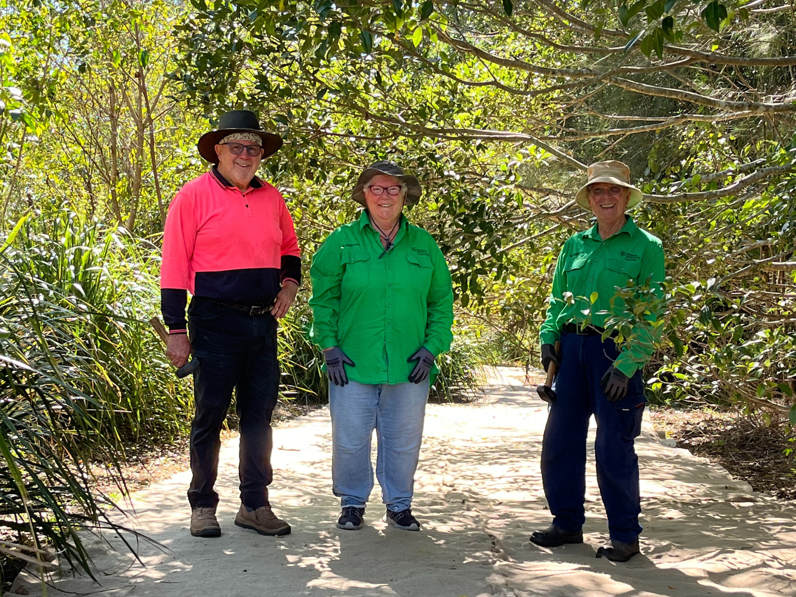 Three Spring Pastures BushCare volunteers, Mary in the centre, smiling at the camera and dressed in their uniforms on-site.