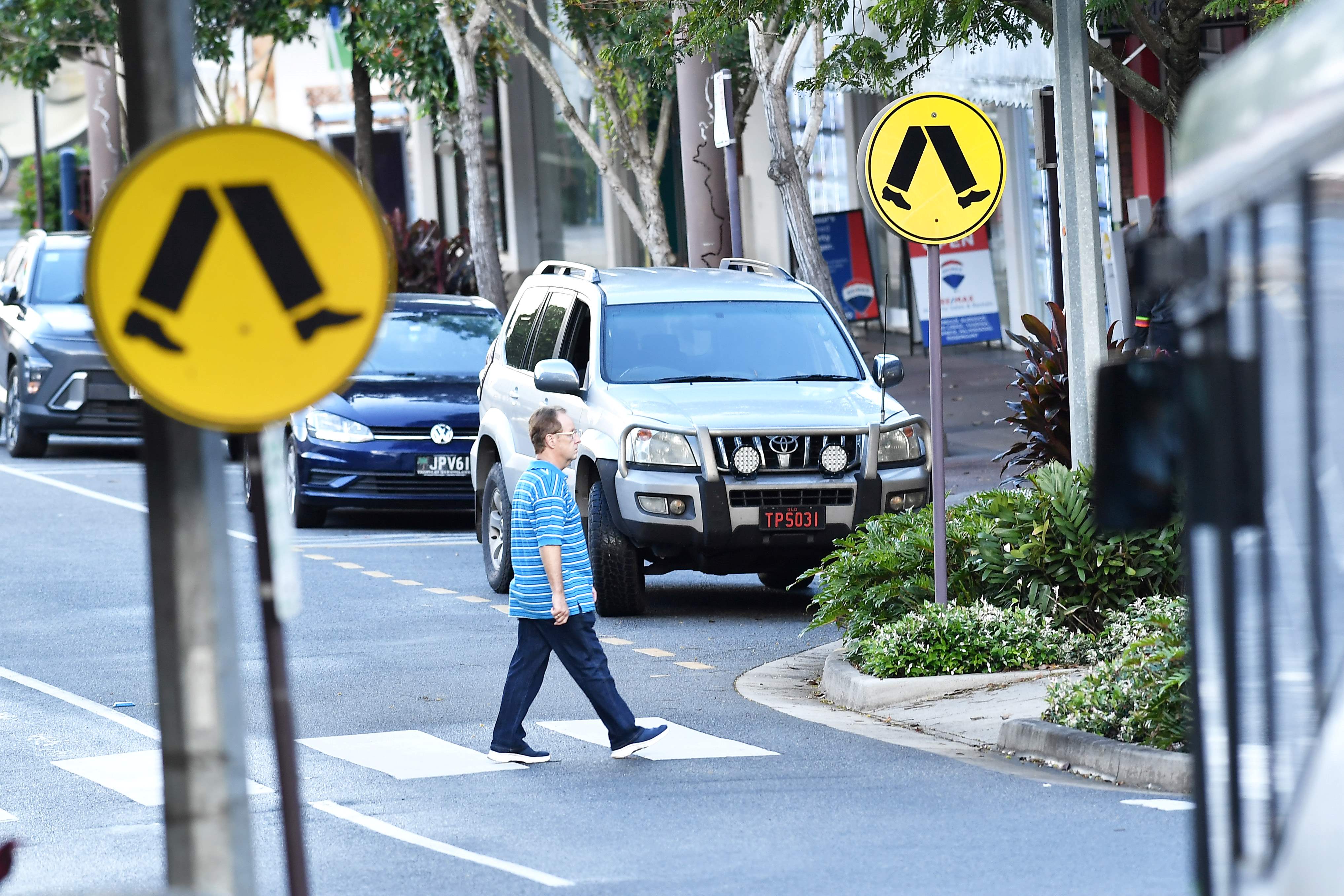 A man walks across a zebra crossing on a busy road. There's a crossing sign in the foreground and background.